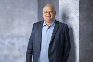 Prof. Dr. Ludger Winkels steht lächelnd vor einer grauen Betonwand und blickt in die Kamera. Er trägt ein dunkelblaues Sakko über einem hellblauen Hemd.