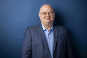 Prof. Dr. Ludger Winkels steht lächelnd vor einem dunkelblauen Hintergrund und blickt in die Kamera. Er trägt ein dunkelblaues Sakko über einem hellblauen Hemd.
